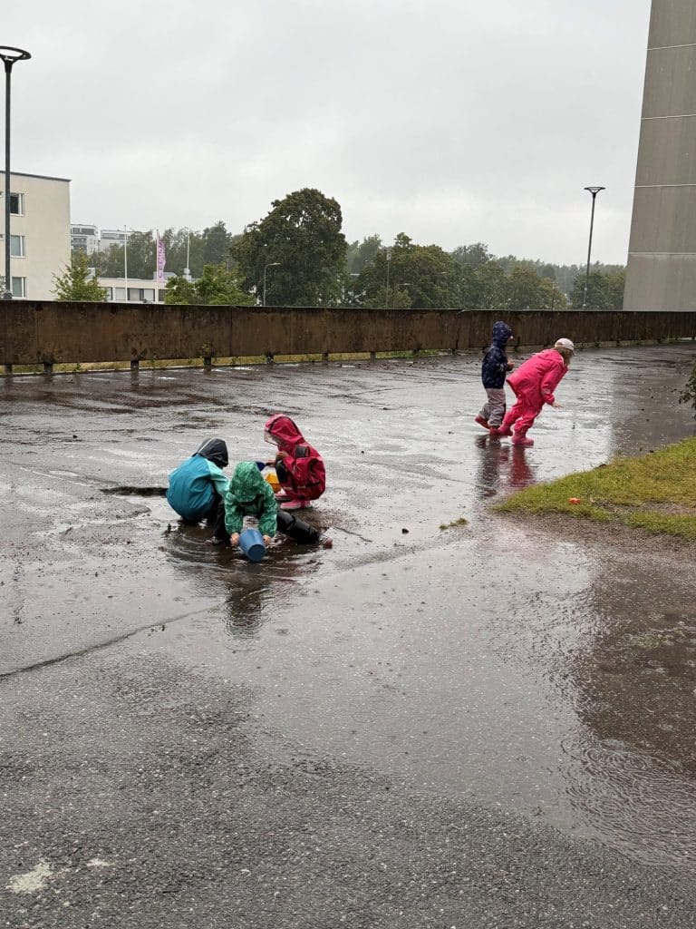 Lapset leikkivät sadevaatteissa lätäköiden äärellä